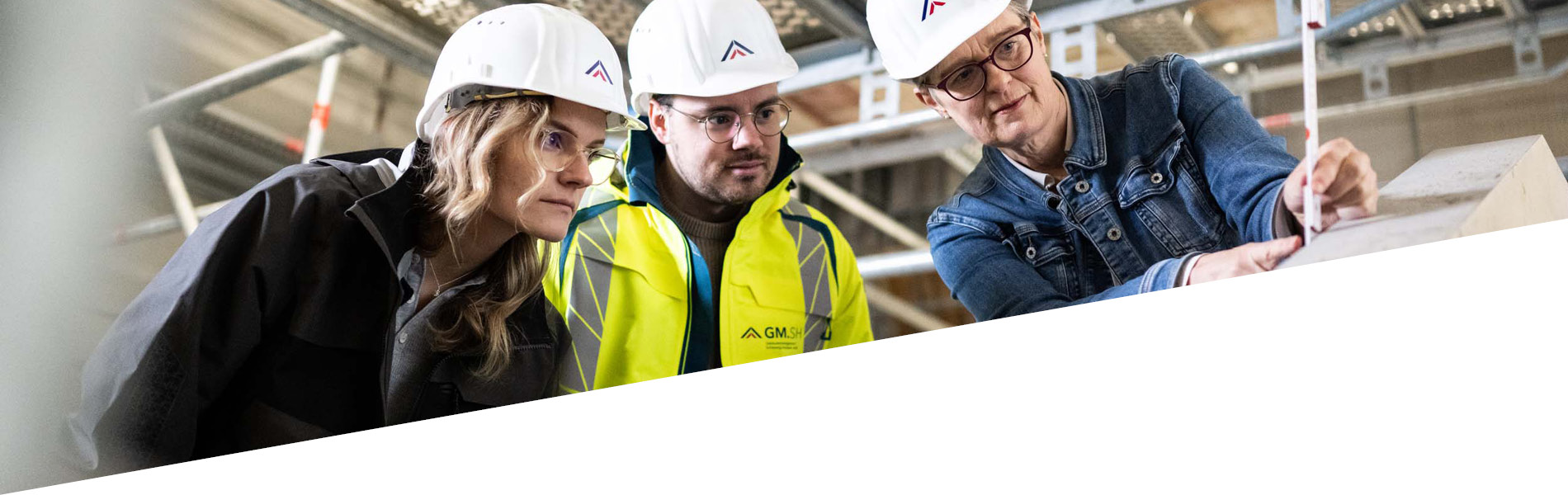 Drei GMSH-Mitarbeiter vermessen etwas auf einer Baustelle. Sie tragen Bauhelme.