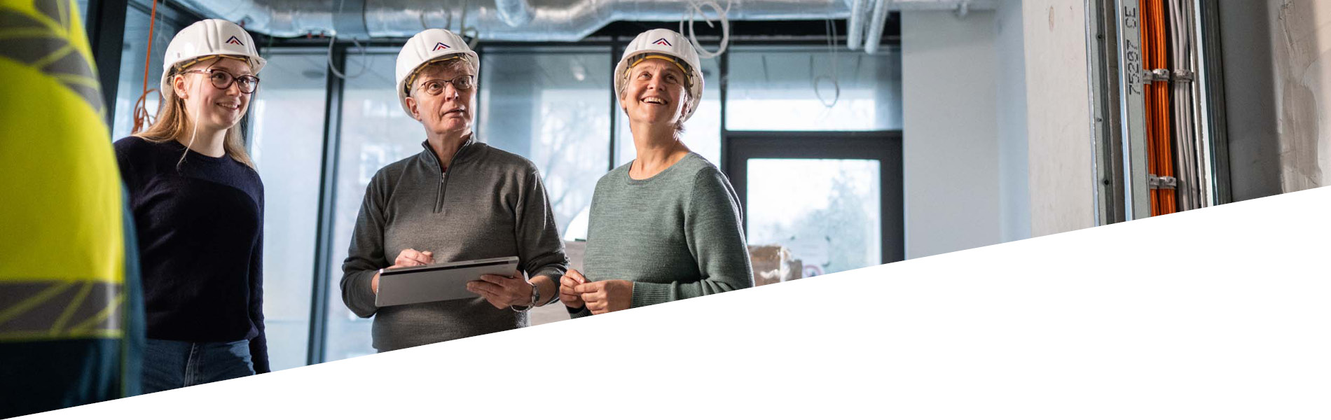 Drei GMSH-Mitarbeiter auf einer Baustelle. Sie tragen Bauhelme. Ein Mitarbeiter hält ein Tablet in der Hand.
