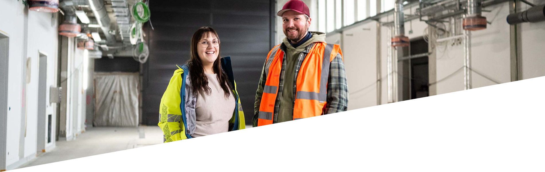 Zwei GMSH-Mitarbeiter lächeln in die Kamera. Sie stehen im Rohbau einer Halle und tragen eine Warnweste.